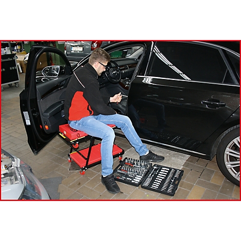 Un homme dans un atelier répare une voiture. Il est assis sur un tabouret rouge et a des boîtes à outils devant lui.